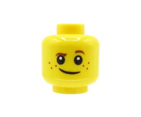 Głowa figurki LEGO w kolorze żółtym, dziecko, piegi, brązowe brwi, uniesiona lewa brew, delikatny uśmiech.